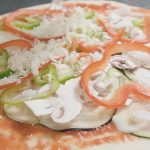 La foto muestra una deliciosa pizza Primavera con una base de tomate natural y una generosa cantidad de queso derretido encima. La pizza está cubierta con una variedad de verduras de temporada, que incluyen pimientos, cebolla y champiñones, dando a la pizza un aspecto colorido y fresco. La masa de la pizza parece estar cruda y apunto de hornear, lo que sugiere que se va a cocinar a la perfección en un horno caliente. En general, la pizza parece ser una opción saludable y deliciosa para cualquier amante de la pizza que quiera disfrutar de una comida sabrosa y nutritiva.